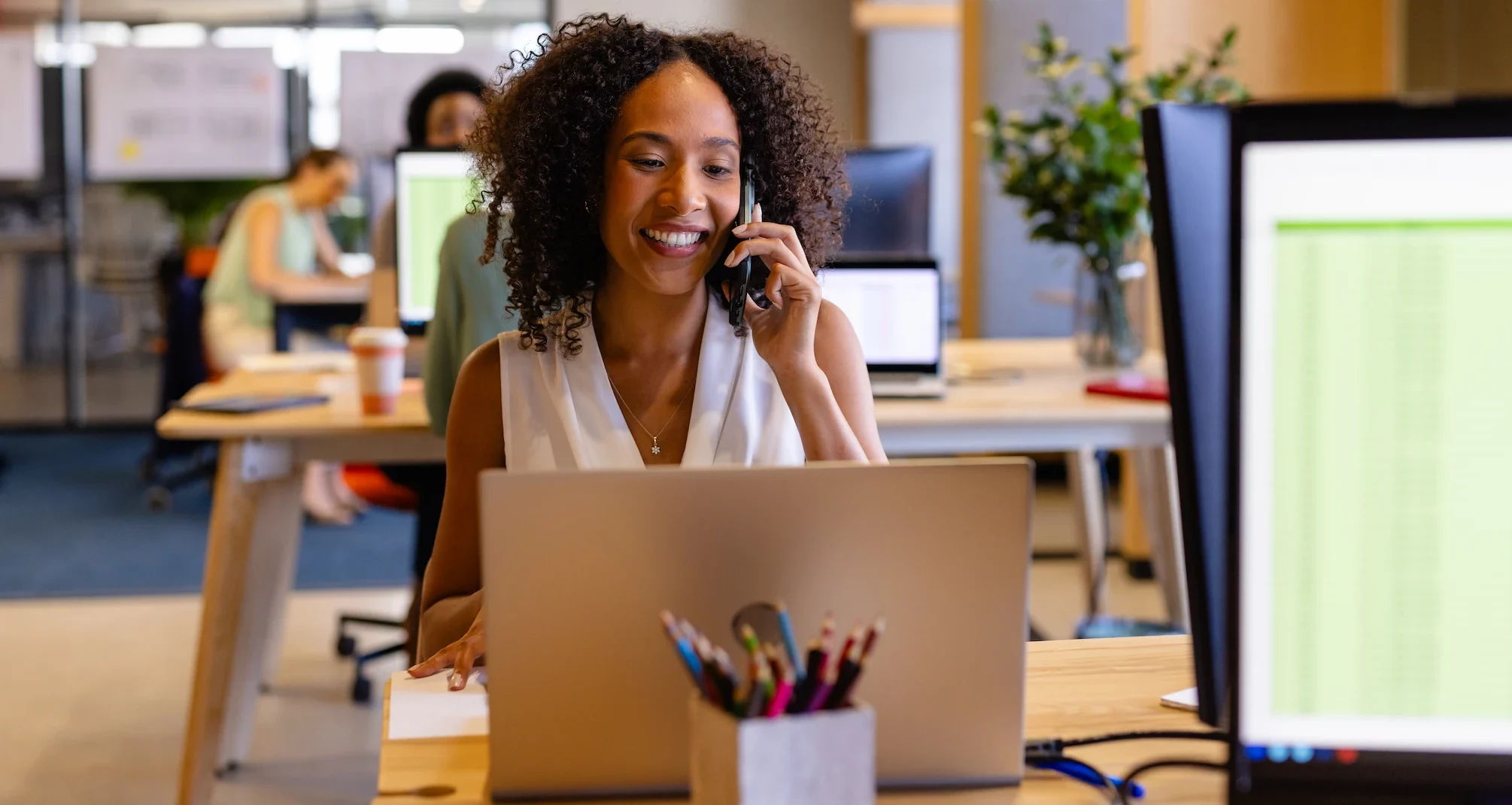 Woman smiling on a phone call at her laptop in a busy open-plan office