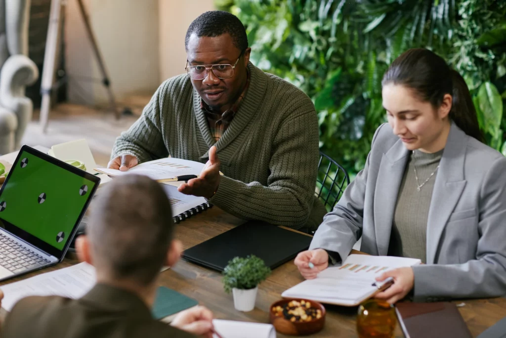 Man gesturing while speaking to colleagues around a table with reports, laptop and a plant